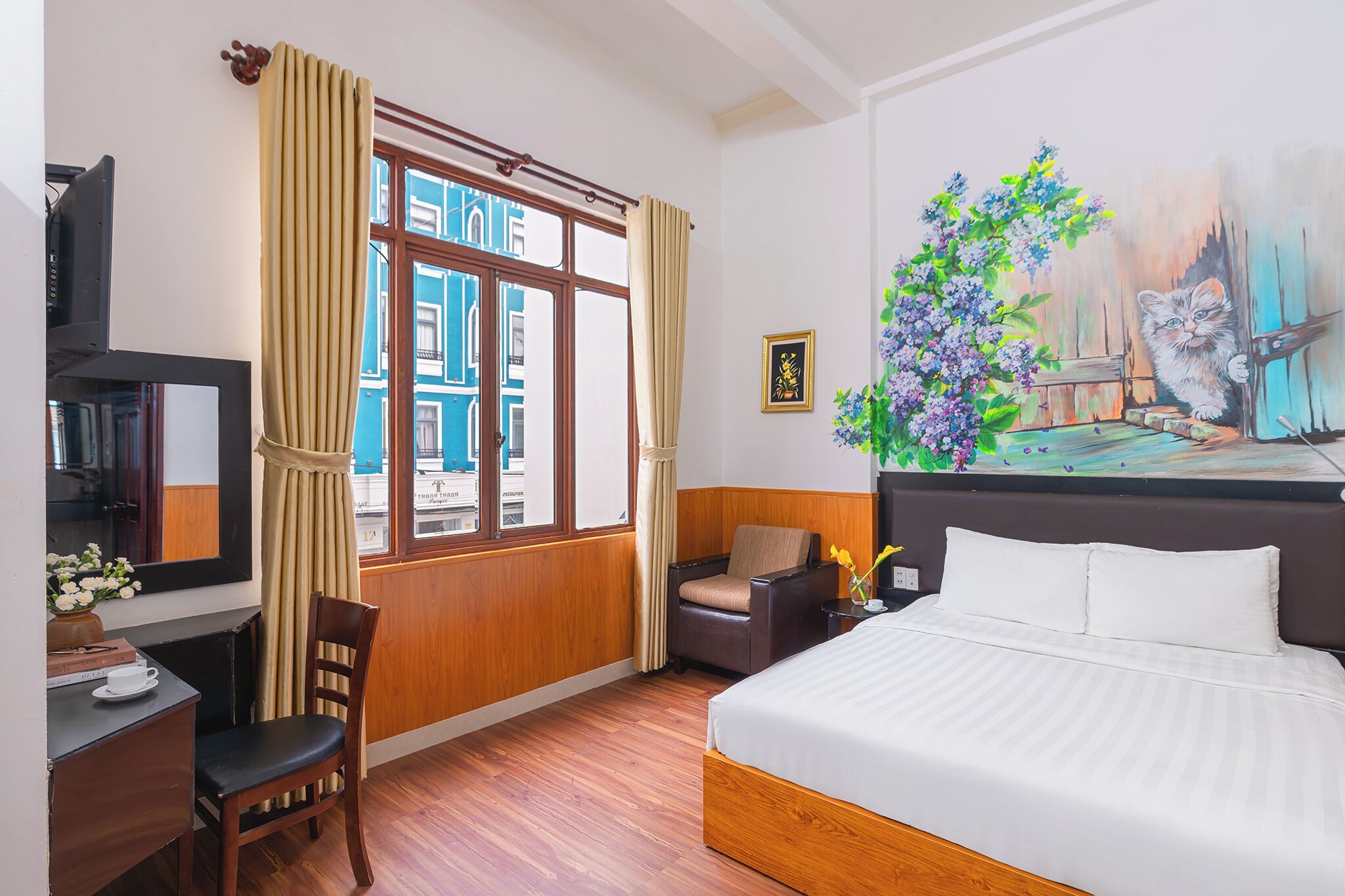 A hotel room with a bed, a desk with a chair, a television, a window with curtains, and a painting on the wall.