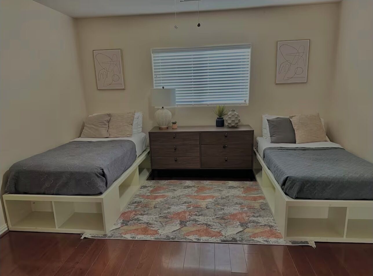 Een slaapkamer met twee eenpersoonsbedden, een kaptafel, een raam met zonwering en een kamerkleed met dessin.
