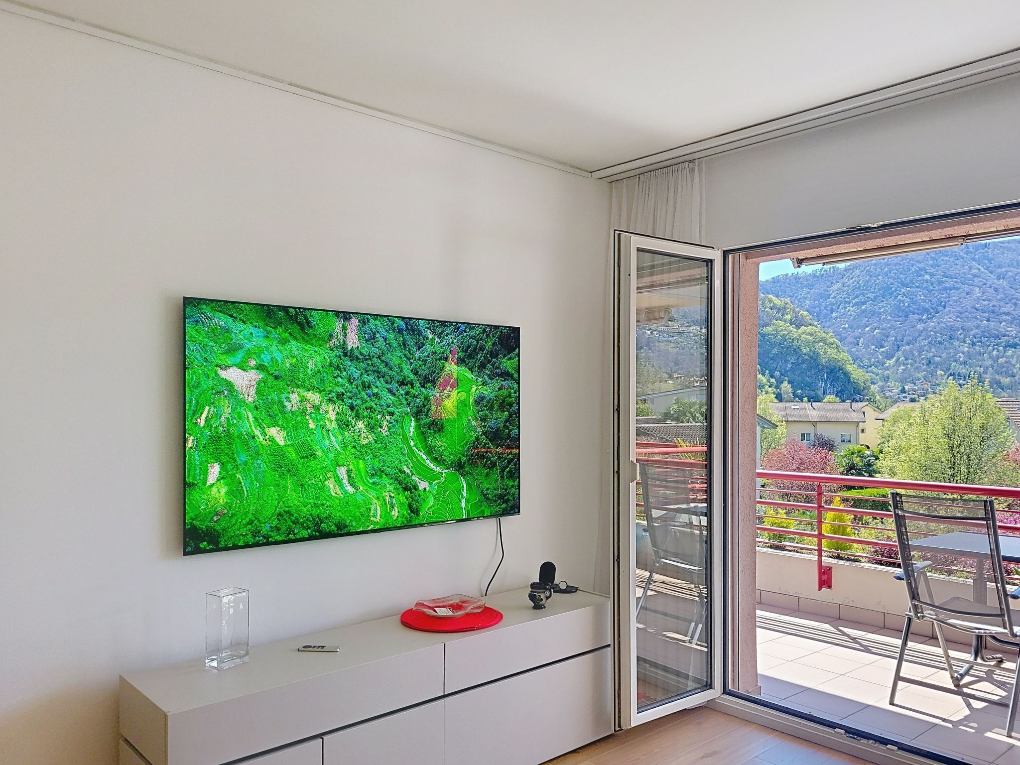 Una habitación moderna con un televisor de pantalla plana grande montado en la pared, una puerta corrediza de vidrio que da a un balcón con vistas a la montaña, y una consola blanca con un objeto rojo encima.