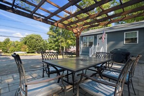 A covered patio with a table and chairs, a grill, and a house in the background. - Near Gunstock | Game Room | Lake Access (Gilford)