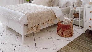 Ein helles Schlafzimmer mit Bett, Nachttischen, einer Pflanze und einem Teppich.
