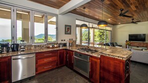 A modern kitchen with wooden cabinets, stainless steel appliances, and large windows offering a view of the outdoors. - Outstanding ocean view, 4 bdr, jungle villa! (Uvita)