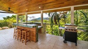 A covered outdoor kitchen area with a wooden ceiling, a bar counter with stools, and a grill. - Outstanding ocean view, 4 bdr, jungle villa! (Uvita)