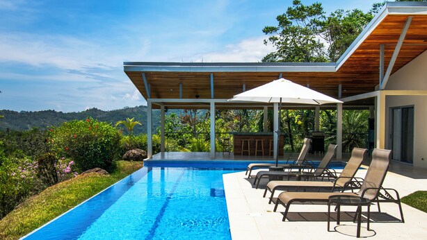A modern outdoor pool area with lounge chairs and a covered seating area, surrounded by lush greenery and a view of distant hills. - Outstanding ocean view, 4 bdr, jungle villa! (Uvita)