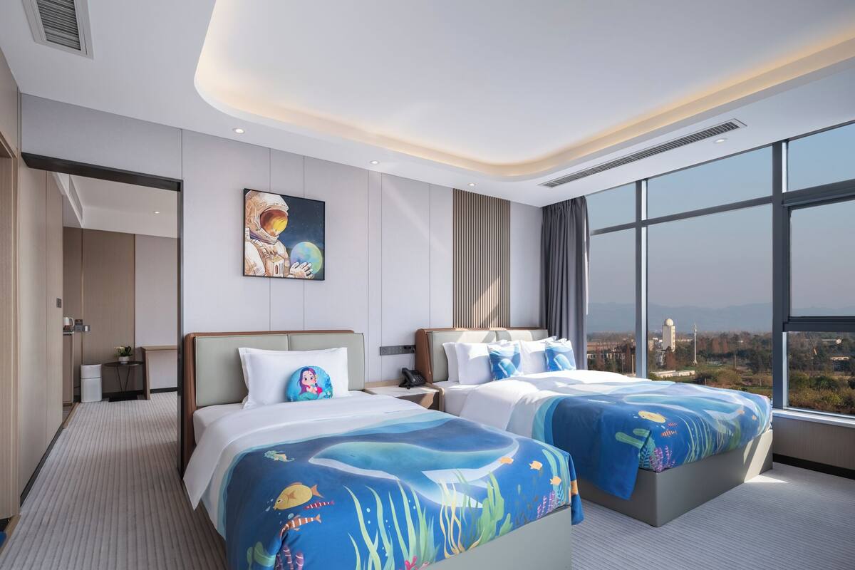 Ein Hotelzimmer mit zwei Betten, einem großen Fenster mit Blick auf die Stadt, einem Bild an der Wand und einem kleinen Tisch mit einer Pflanze.