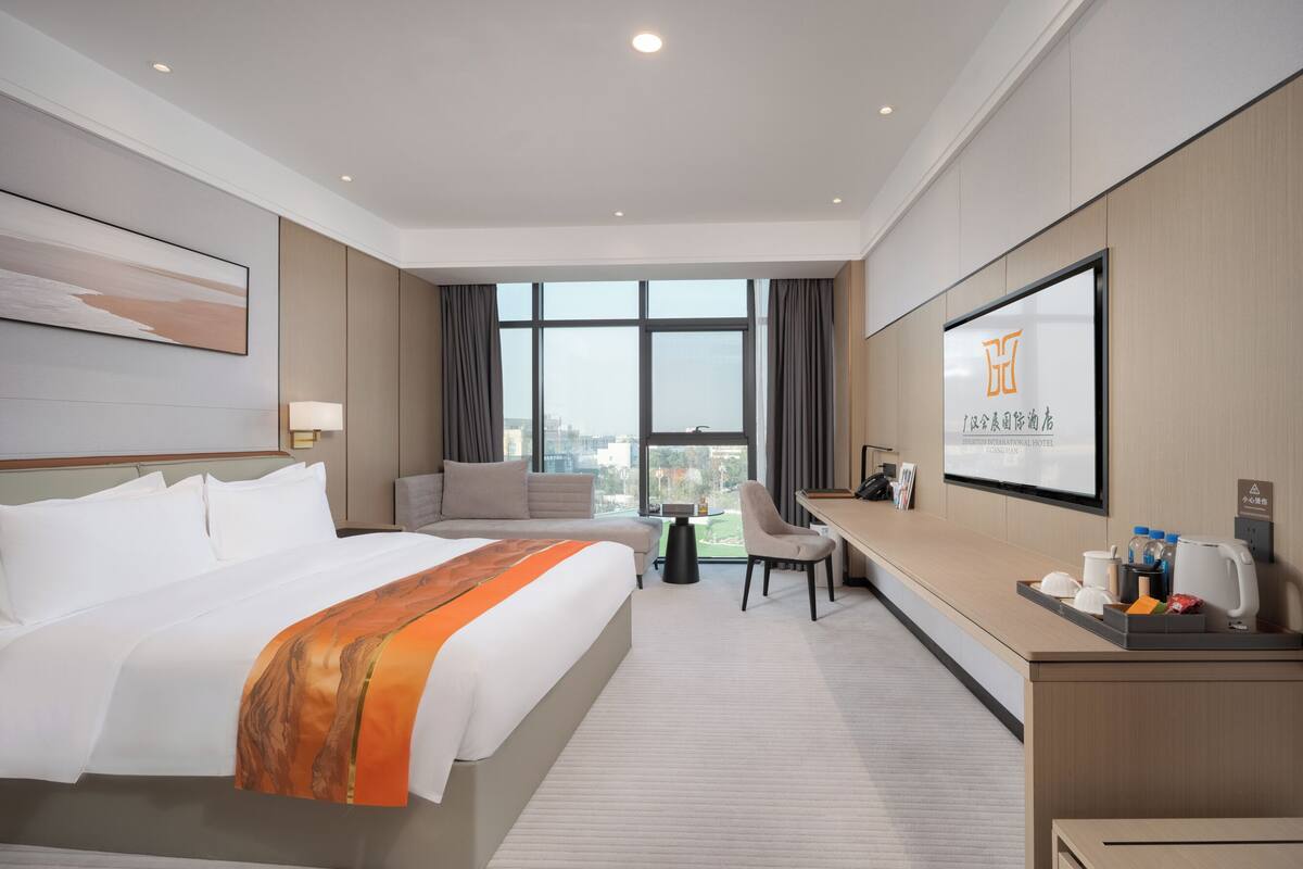 Ein Hotelzimmer mit einem großen Bett, einem Schreibtisch, einem Sessel und einem Fernseher, der Hotelinformationen anzeigt.