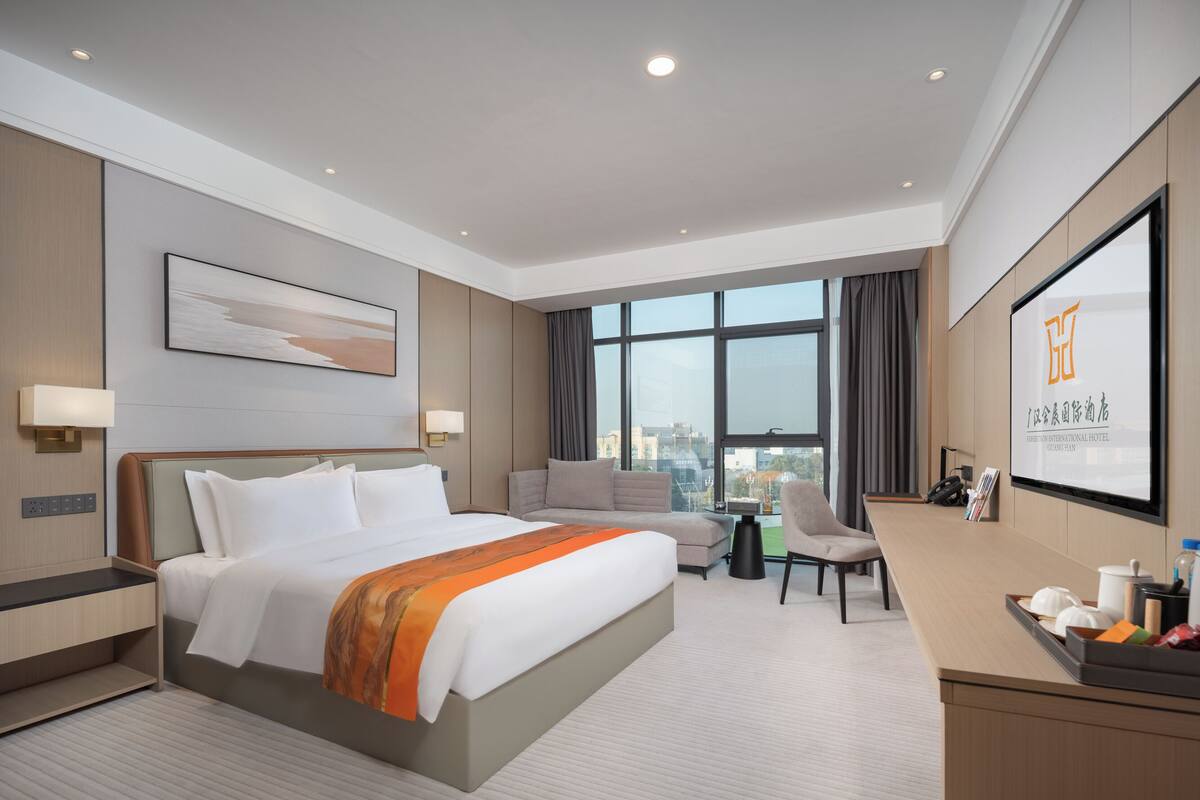Ein modernes Hotelzimmer mit einem großen Bett, einem Schreibtisch, einem Sessel und Blick auf die Stadt durch große Fenster.