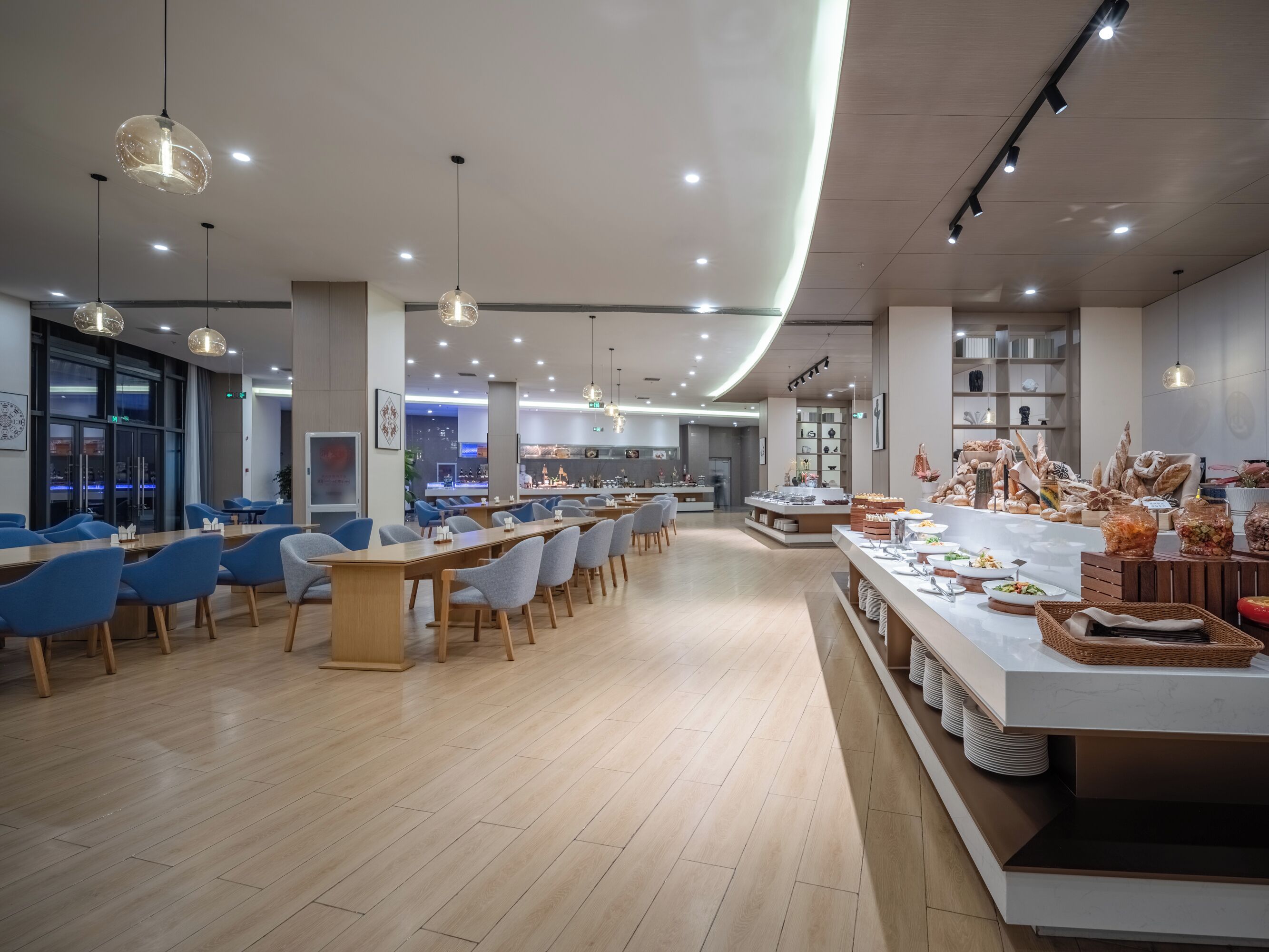 A modern dining area with long tables and blue chairs, a buffet counter with various food items, and a kitchen in the background.