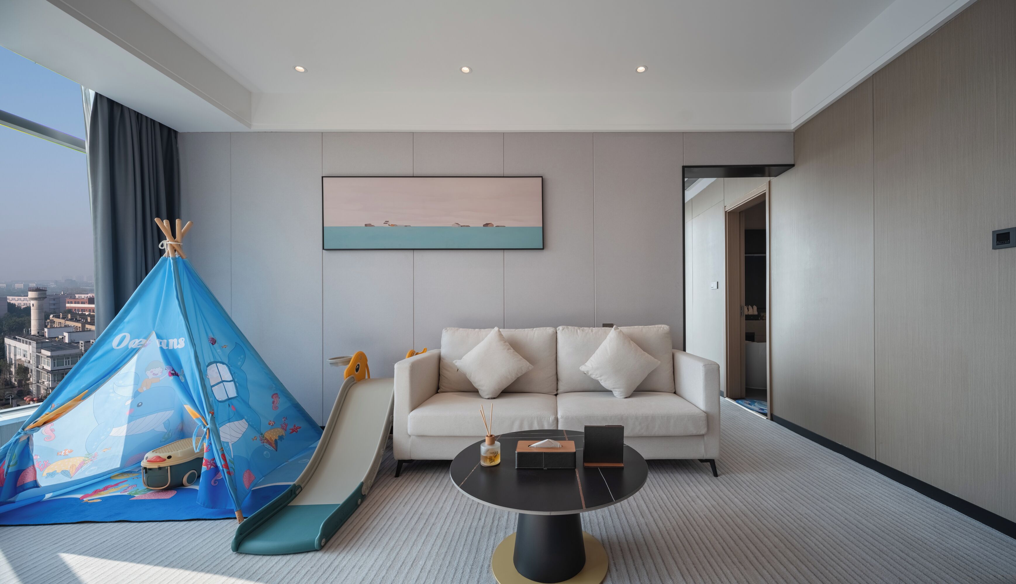 Una habitación de hotel moderna con un sofá blanco, una mesita de centro negra redonda y un área de juegos azul con forma de tienda de campaña, llena de juguetes.