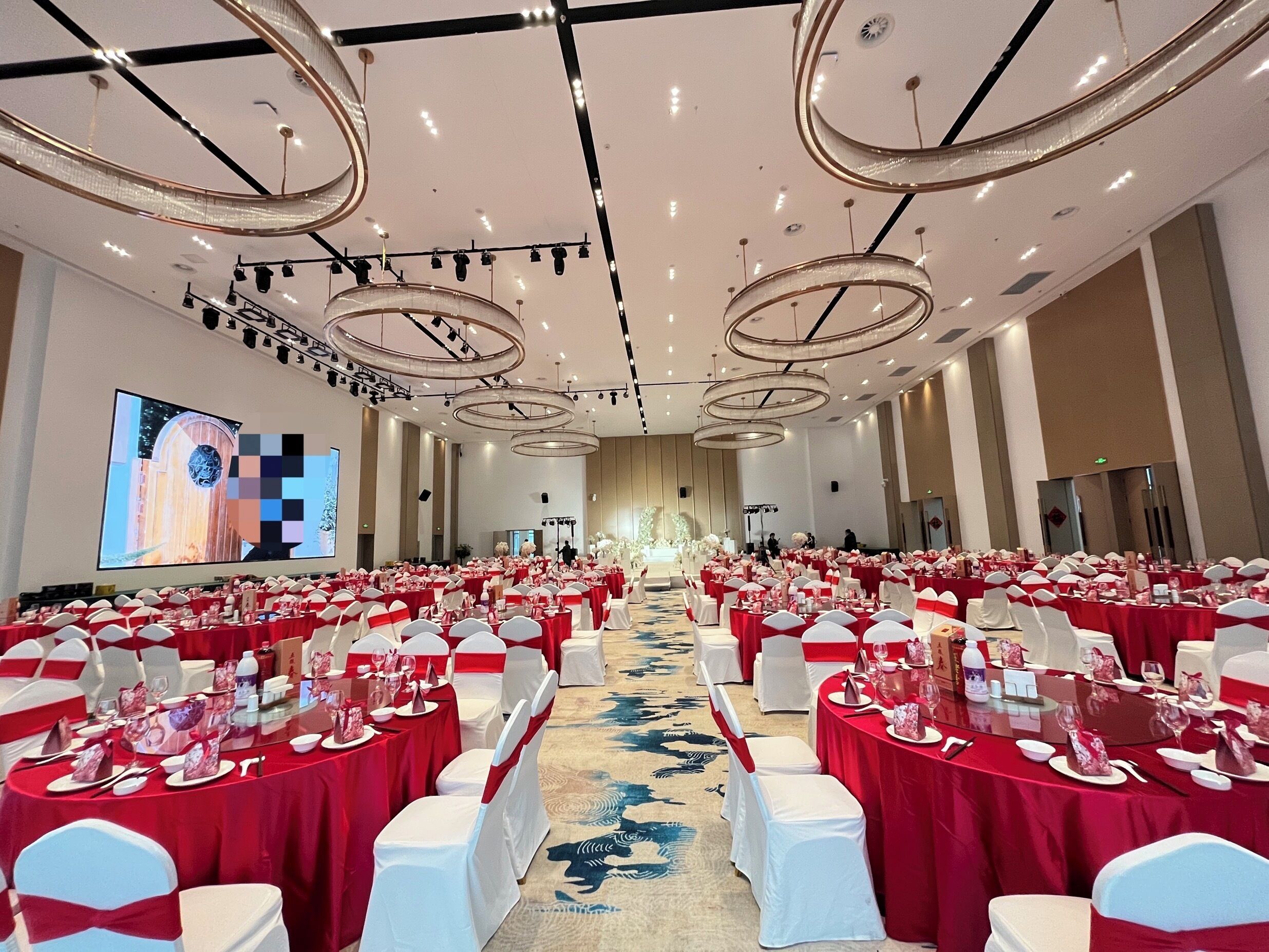 A large banquet hall with round tables covered in red tablecloths and white chairs, a large screen on the wall, and a carpeted floor.