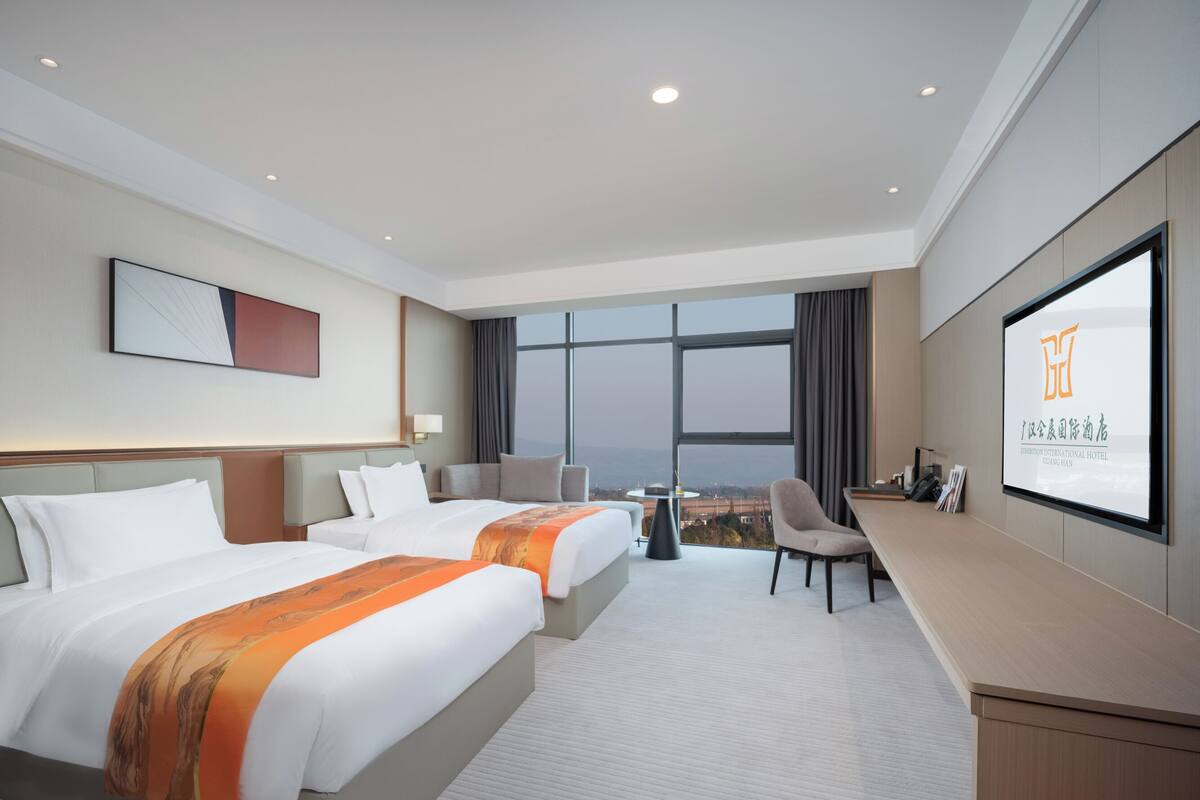 Ein Hotelzimmer mit zwei Betten, einem Schreibtisch, einem Stuhl, einem Fernsehbildschirm und einem großen Fenster mit Blick auf die Stadt.