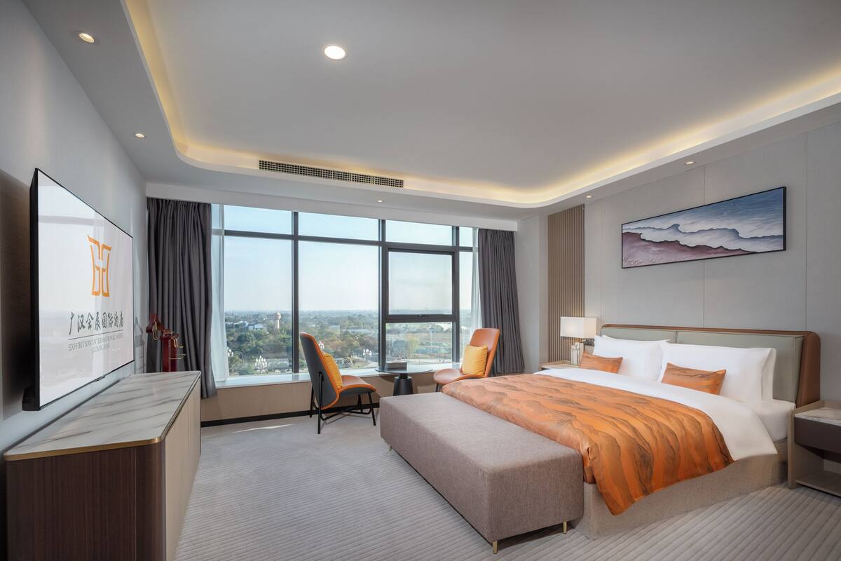 Ein modernes Hotelzimmer mit einem großen Bett, einem Schreibtisch, einem Sessel und einem Fernseher, der ein Logo zeigt.