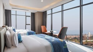 Una habitación de hotel moderna con una cama grande, dos sillas, una mesita y vistas a la ciudad a través de amplios ventanales.