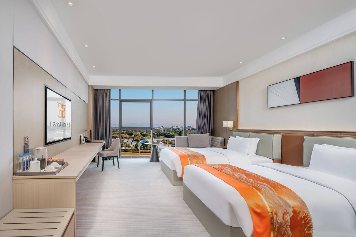 Ein Hotelzimmer mit großem Fenster, Schreibtisch, zwei Betten und Blick auf die Stadt.