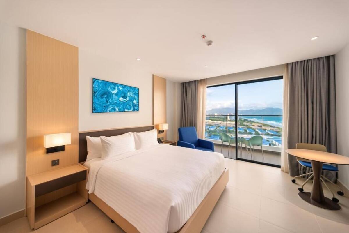 A hotel room with a large bed, a desk, a chair, a view of the sea, and a blue artwork on the wall.