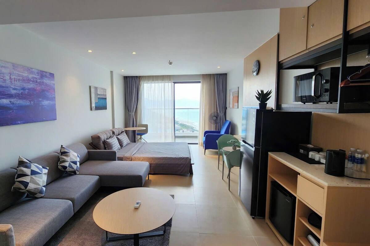 A modern living room with a sofa, a round coffee table, a dining area with a table and chairs, a kitchen with appliances, and a large window offering a view.