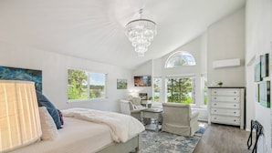 A bedroom with a large bed, a chandelier, a desk, and a chair. There are windows with views of greenery. - Lake Front Home, Newly Remodeled, Hot Tub, Game Room, Views! (Pagosa Springs)
