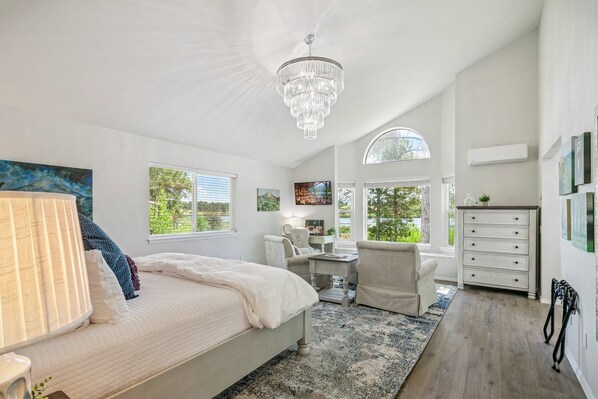 A bedroom with a large bed, a chandelier, a desk, and a chair. There are windows with views of greenery.