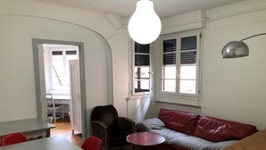 A room with a wooden floor, a red sofa, a small round table, a dark armchair, a white table with red chairs, and a window with blinds. - Family apartment near Lot (CAHORS)