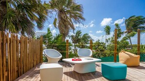 Una terraza de madera con una mesa redonda y dos sillas blancas, un otoman turquesa y un cojín color beige.