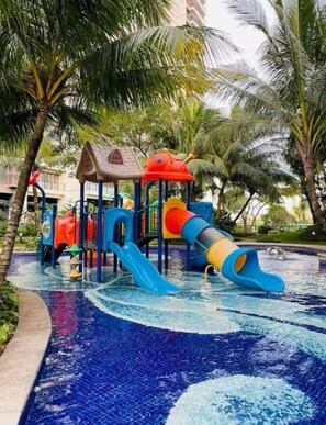 Un parque infantil con toboganes y estructuras para escalar, rodeado de palmeras y una fuente de agua.