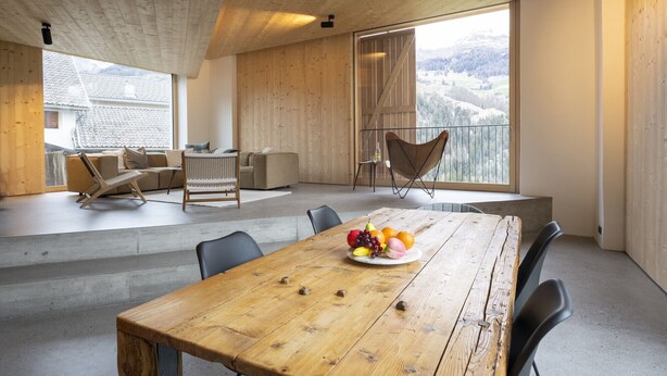 Ein geräumiger Raum mit Holzbalkendecke, einem großen Esstisch mit Obst, einer Couch und einem Sessel.