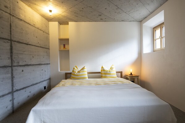 A bedroom with a bed, pillows, a nightstand, a lamp, a window, and a shelf with books.