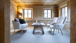 Ein gemütliches Blockhaus-Interieur mit Holzbalkendecke, zwei weißen Sesseln, einem kleinen Holztisch und einer Couch mit Kissen.