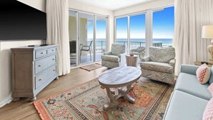 A spacious living room with a large window offering a view of the ocean, a balcony with outdoor seating, and a traditional wooden coffee table. - The Crescent 306 (Miramar beach)