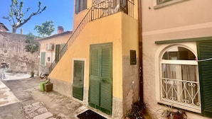 Un edificio con una escalera que conduce a un balcón, contraventanas verdes y un suelo de piedra.