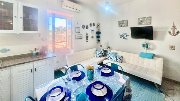 Una habitación con una mesa de comedor preparada para cuatro personas, un sofá blanco, un televisor montado en la pared y una ventana con vistas a edificios.