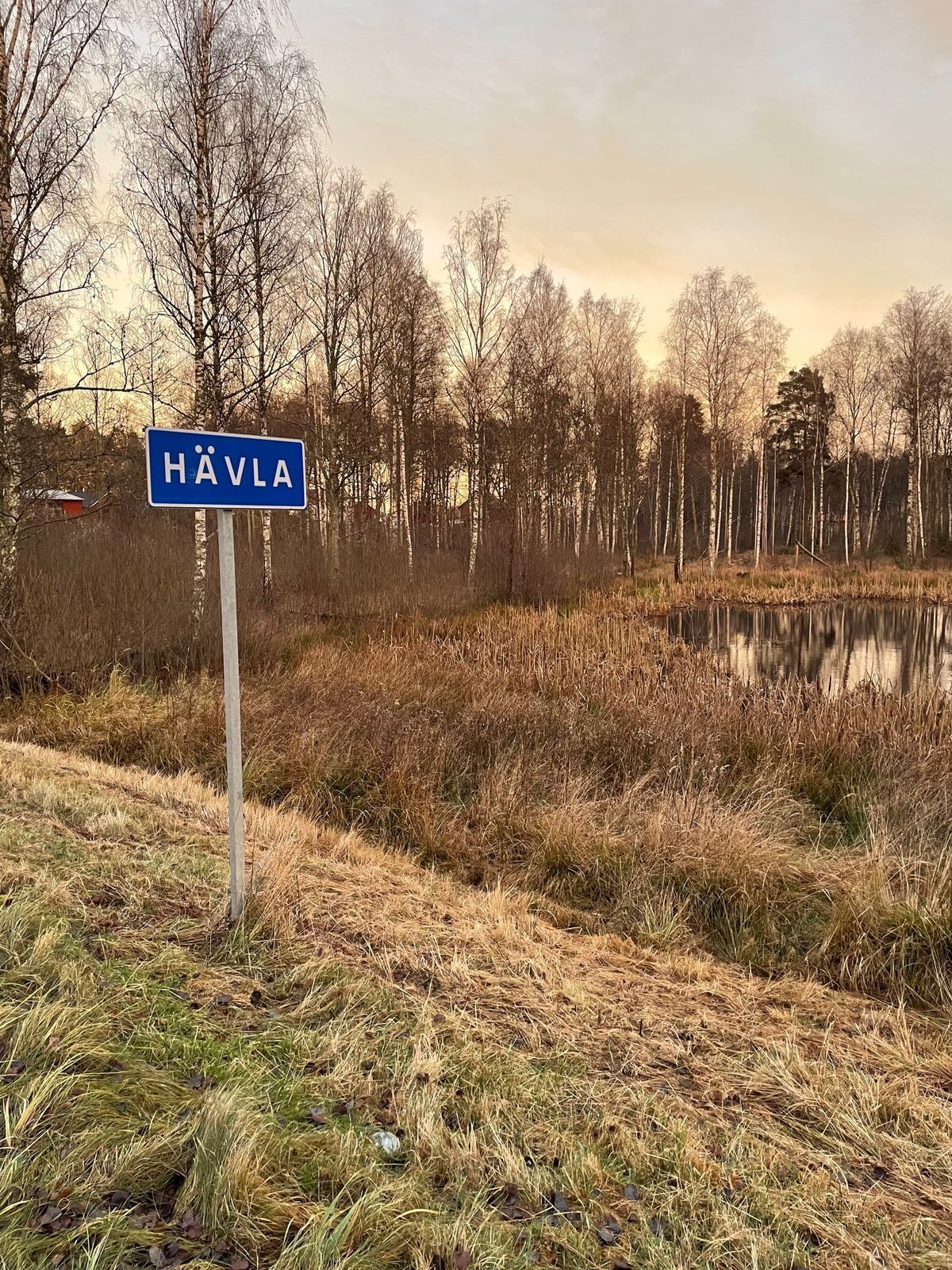 Uma placa com o nome 'HÄVLA' em ambiente rural, com um corpo d'água e árvores ao fundo.