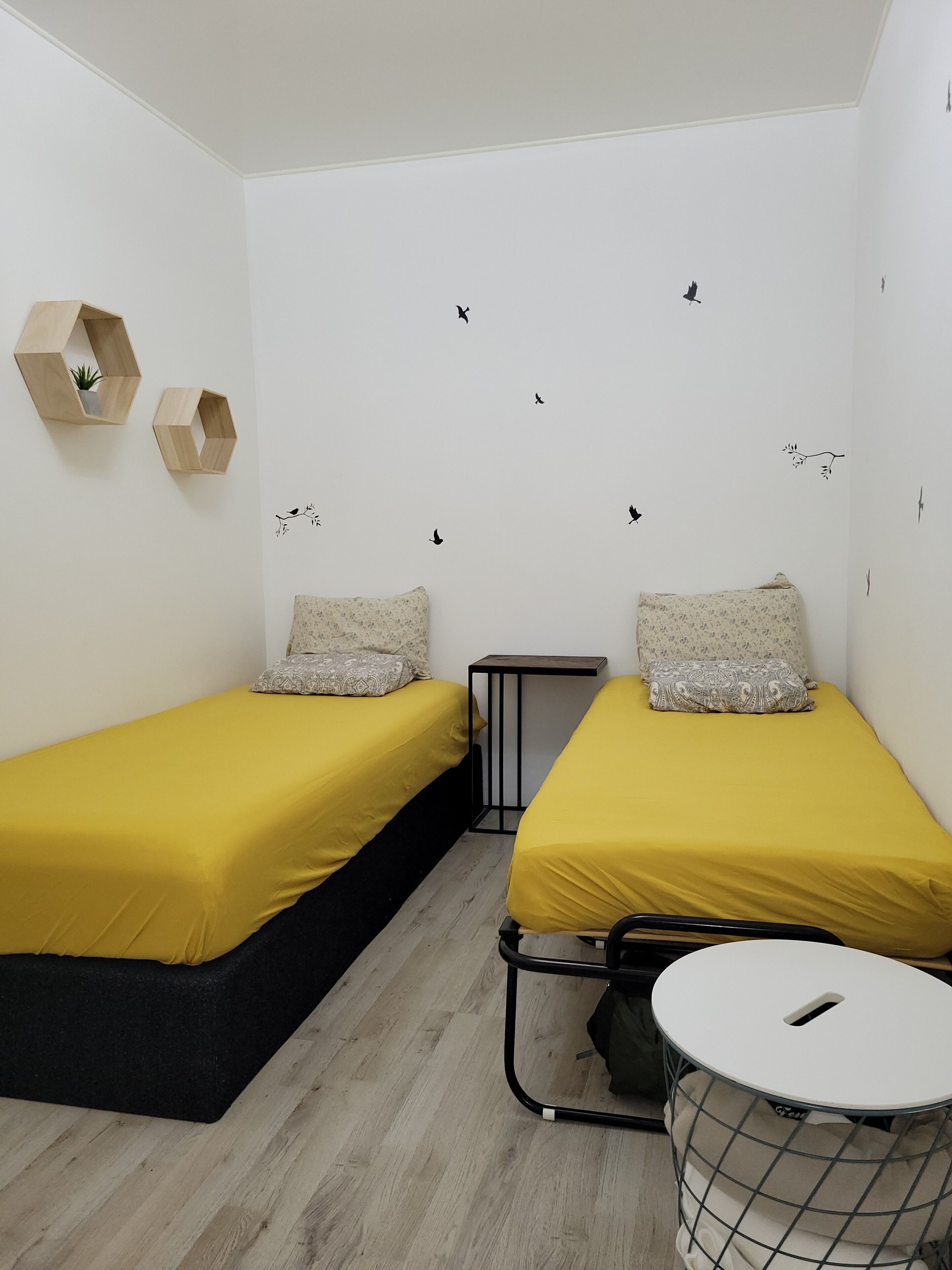 Een lichte, moderne kamer met twee bedden, een tafeltje en een rond opbergmeubel.