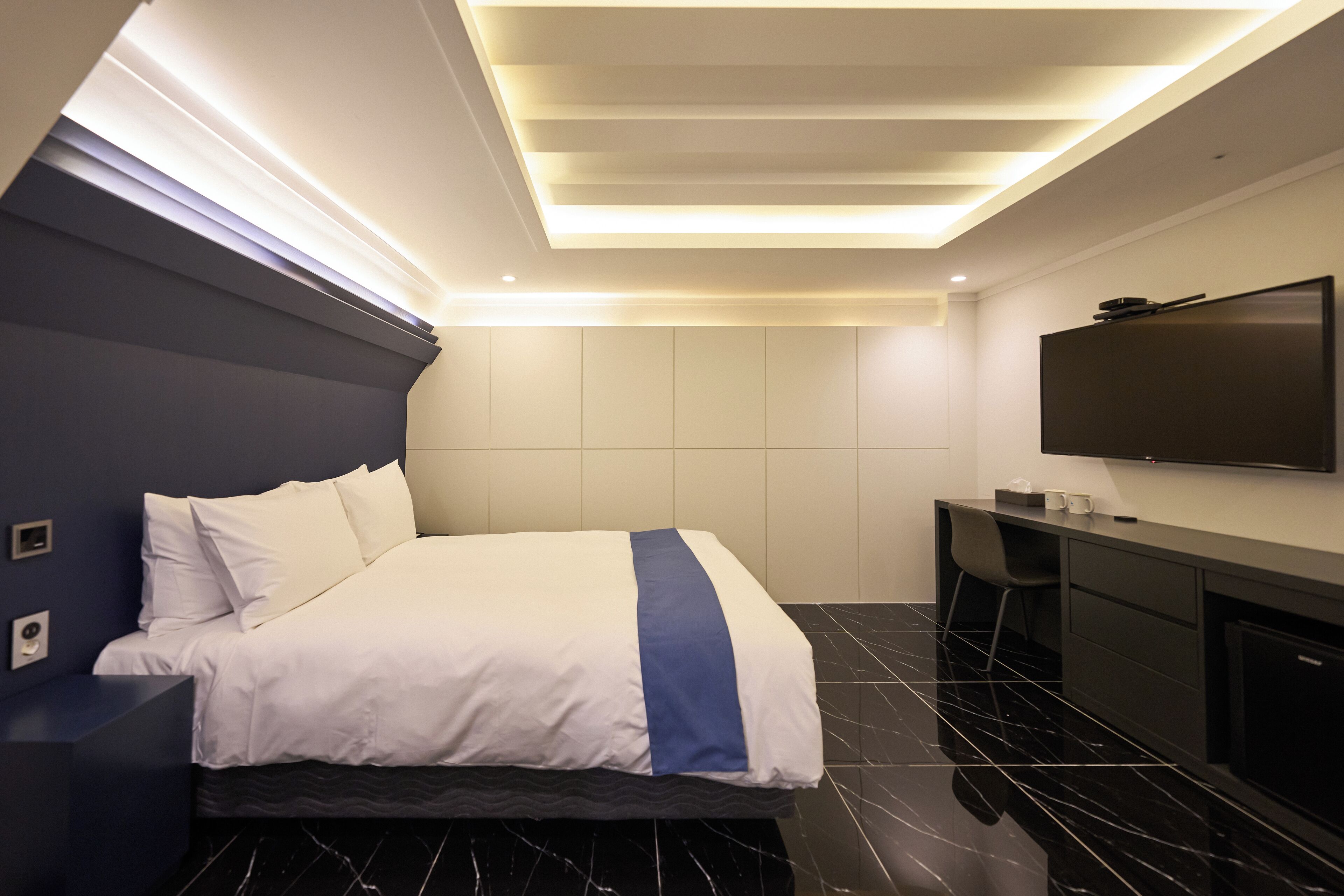 Quarto de hotel moderno com cama, escrivaninha, TV de tela plana e teto com iluminação embutida.