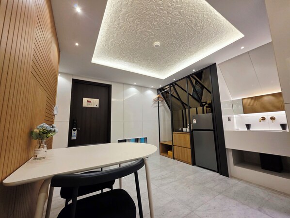 A modern hotel room with a white table, black chairs, a wooden accent wall, a decorative ceiling, and a kitchen area with a refrigerator and wooden cabinets. - BlueDuck Hotel (Seoul)
