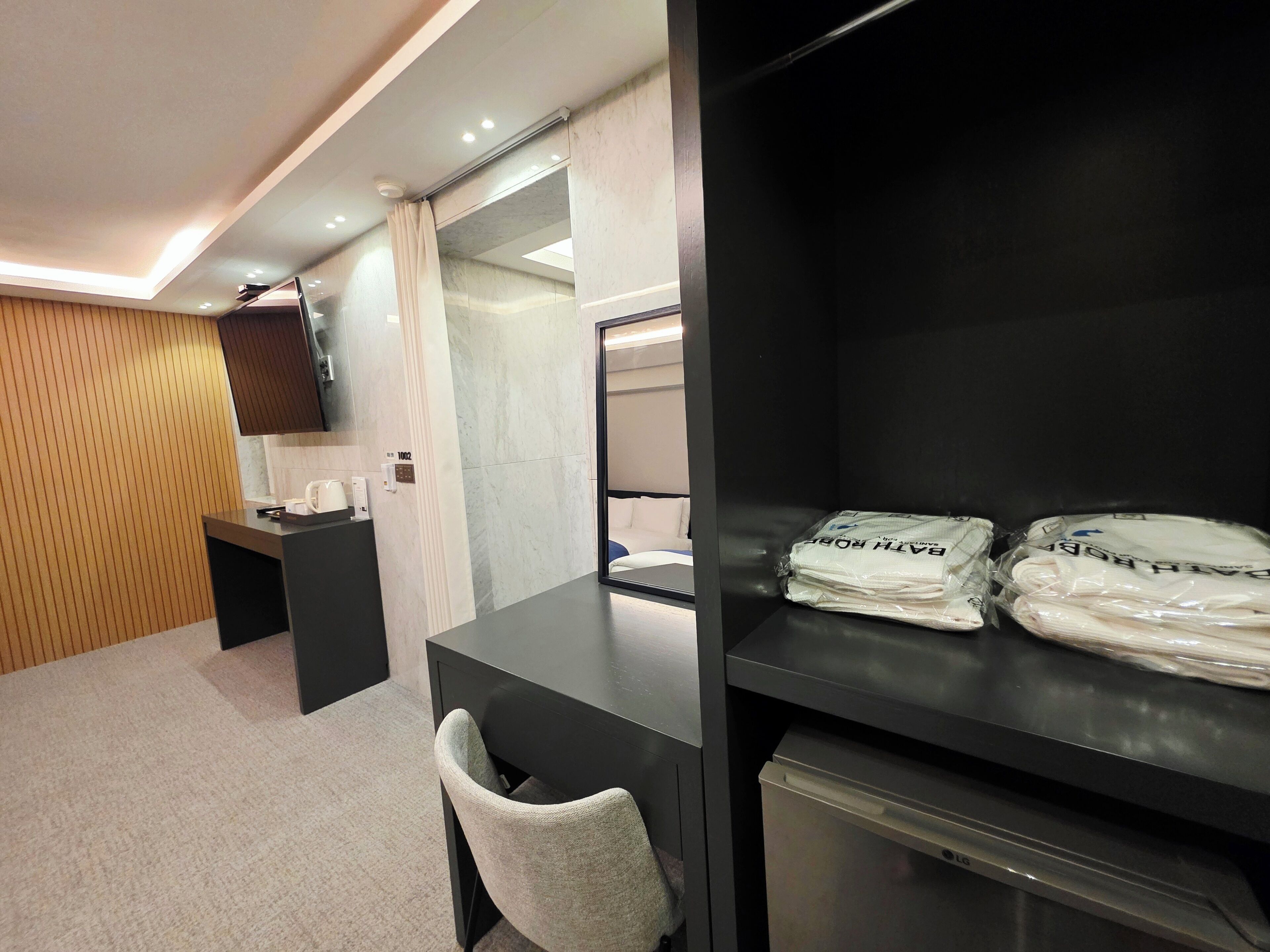 A hotel room with a desk, a chair, a bed, a TV mounted on the wall, and a shelf with folded towels.