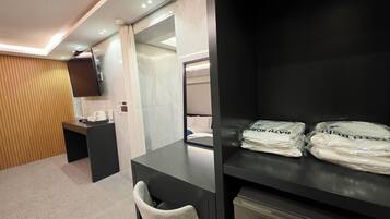 A hotel room with a desk, a chair, a bed, a TV mounted on the wall, and a shelf with folded towels.