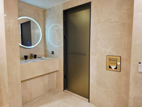 Un baño moderno con tocador, espejo con iluminación circular, puerta y cubo de basura.