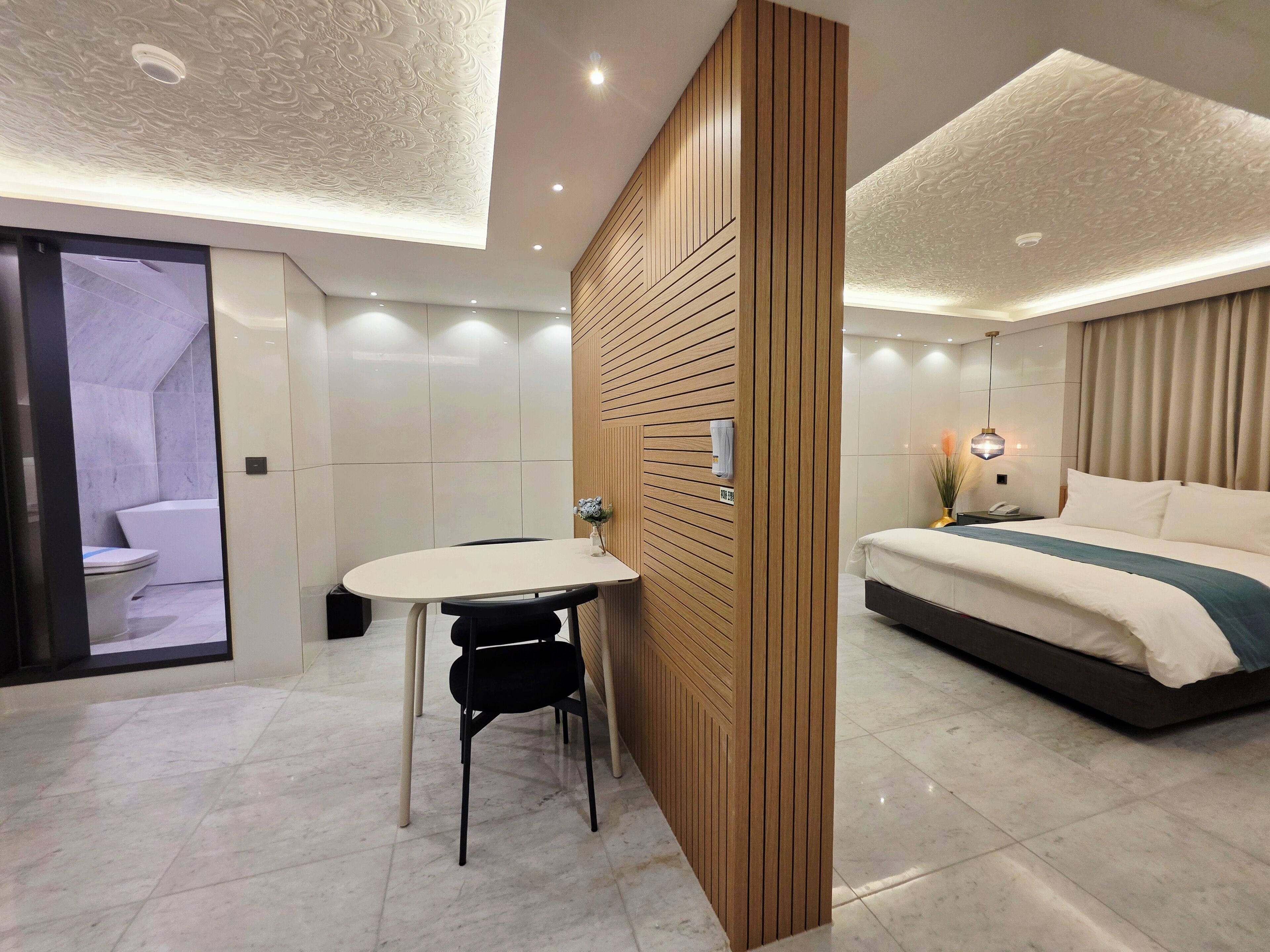 A modern hotel room with a wooden partition, a bed, a bedside table, and a bathroom visible through an open door.