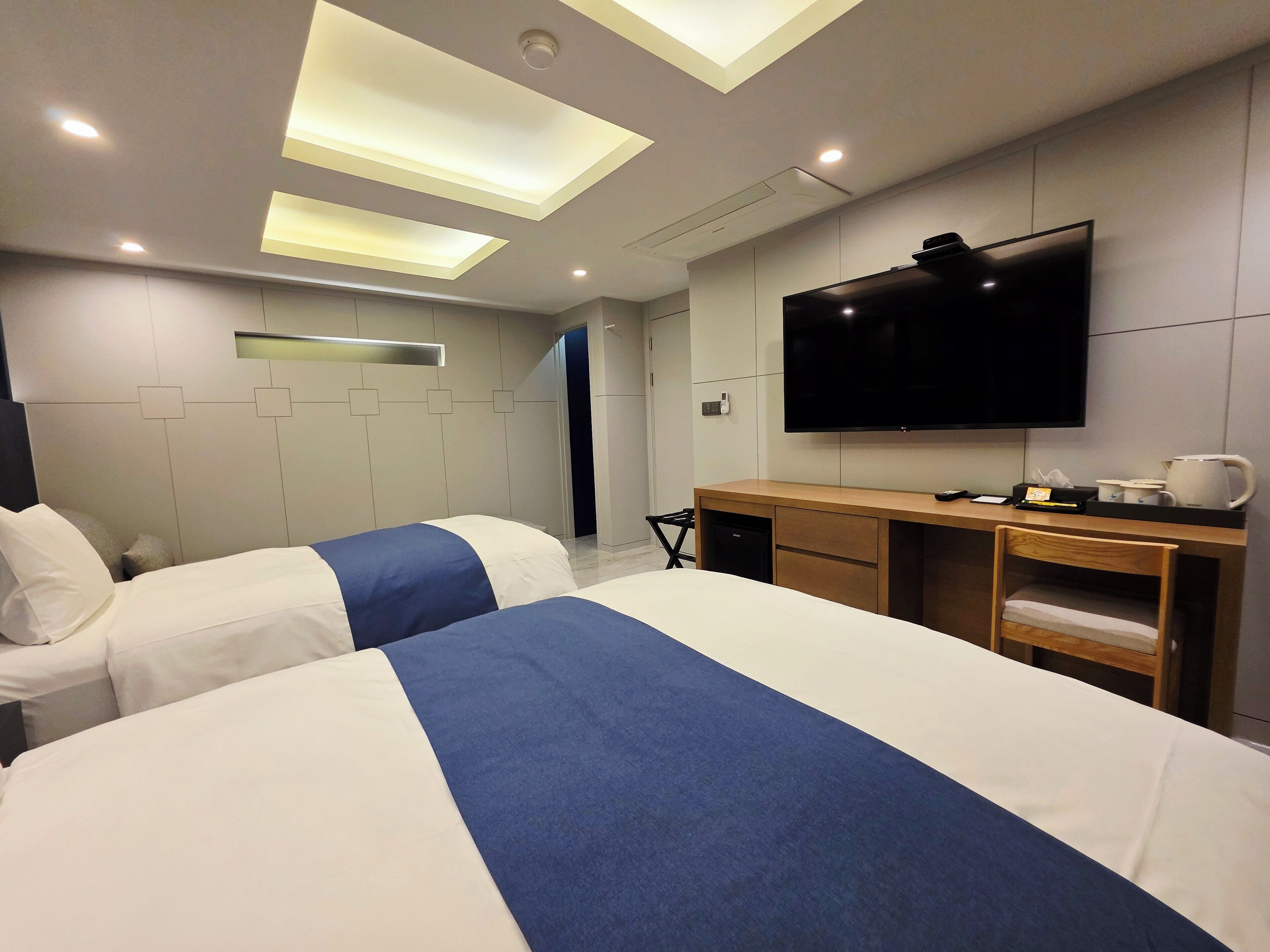 A modern hotel room with two beds, a flat-screen TV, a wooden desk, and a ceiling with recessed lighting.