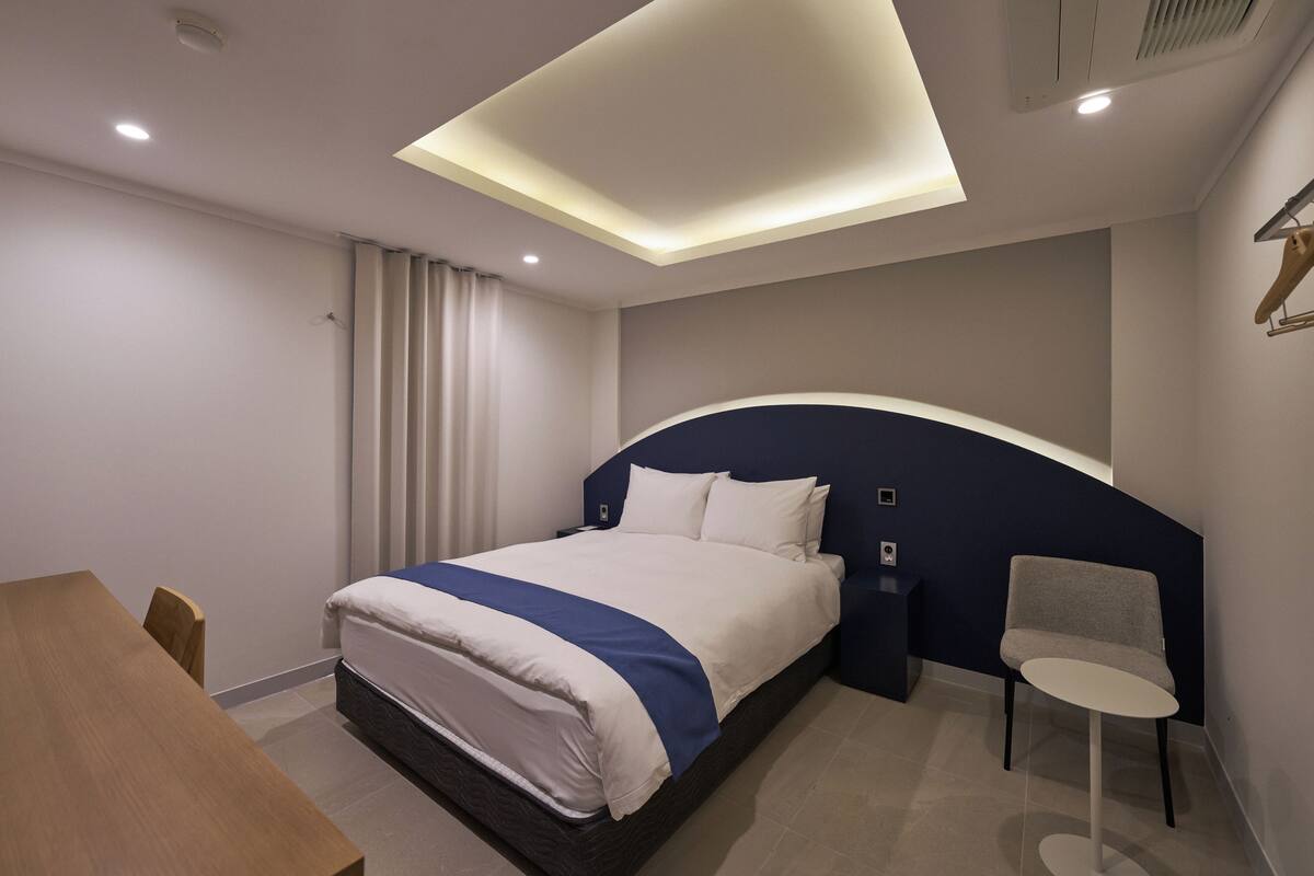 A modern hotel room with a large bed, a small table, a chair, and a ceiling light fixture.