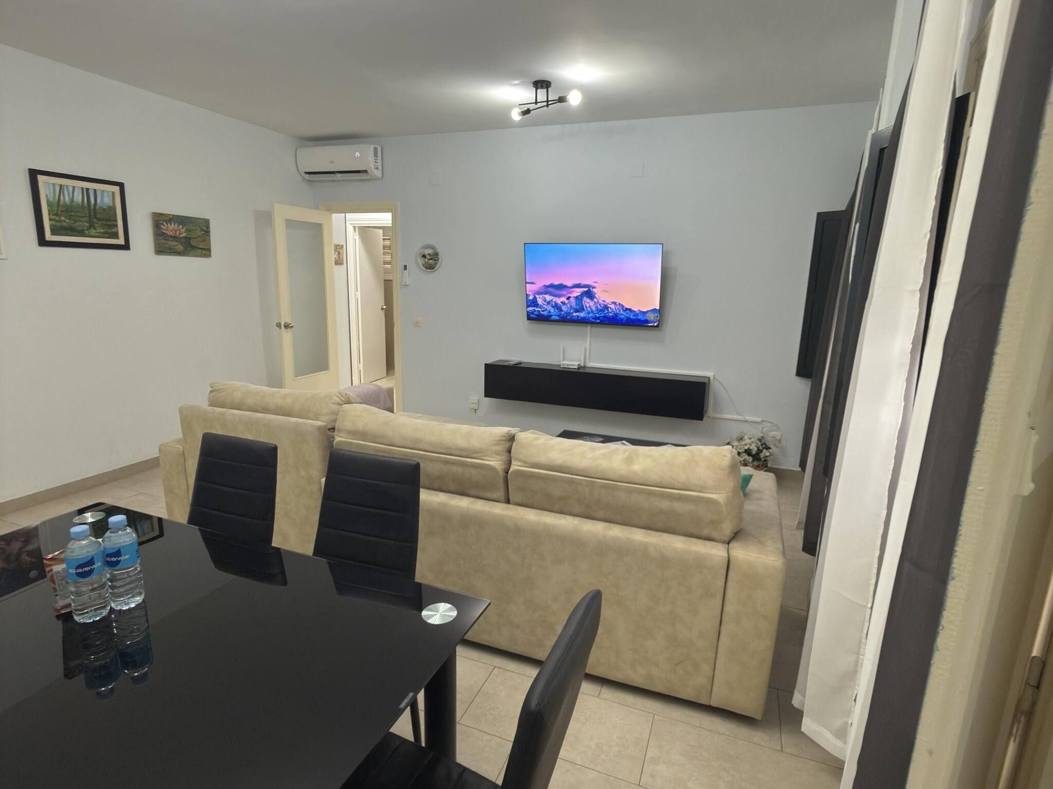 Una sala de estar con un sofá beige, un televisor de pantalla plana montado en la pared, una mesa de comedor con sillas y un equipo de aire acondicionado.