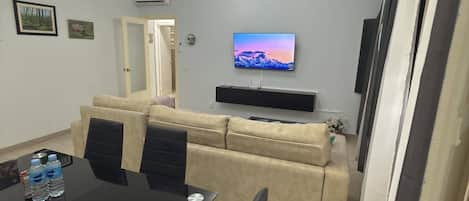 A living room with a beige sofa, a flat-screen TV mounted on the wall, a dining table with chairs, and an air conditioning unit.