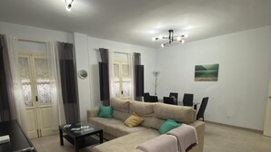 Ein Wohnzimmer mit einem beigefarbenen Sofa, einem Couchtisch und einer TV-Konsole. An den Fenstern hängen Vorhänge, an der Wand hängt ein Gemälde und eine Chandelieber ist angebracht.