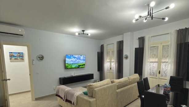 Ein modernes Wohnzimmer mit einem beigen Sofa, einem wandmontierten Flachbildfernseher, einem Esstisch mit Stühlen und einer Deckenleuchte.
