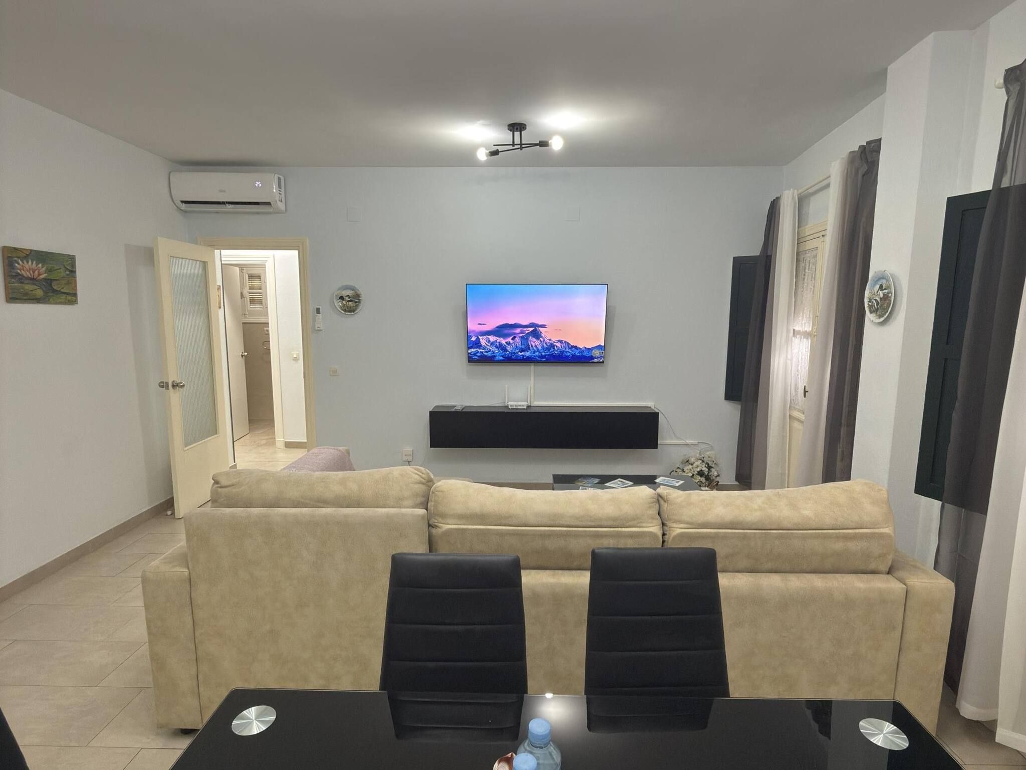 Ein Wohnzimmer mit einem beigefarbenen Sofa, einem Wand-Fernseher, einer Deckenleuchte und einer Tür zu einem anderen Raum.