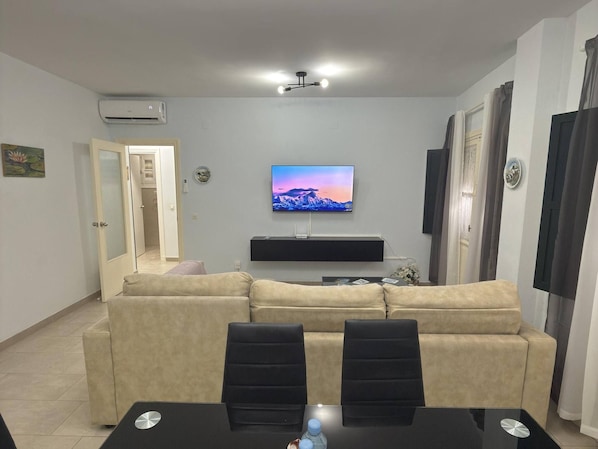 A living room with a beige sofa, a flat-screen TV mounted on the wall, a ceiling light fixture, and a door leading to another room. - García Rota Beach by staycare (Rota)