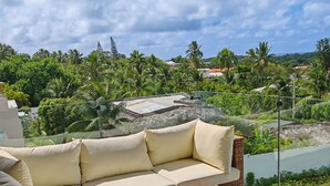A modern outdoor seating area with a sofa and wicker chairs, overlooking a lush landscape with palm trees and a river. - Modern Seaview Condo w/ Pool & Balcony – Steps to Beach (Cabarete)