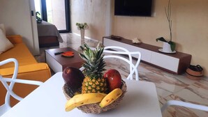 A modern living room with a white sofa, a dining table with a fruit basket, a TV mounted on the wall, and a potted plant. - Modern Seaview Condo w/ Pool & Balcony – Steps to Beach (Cabarete)