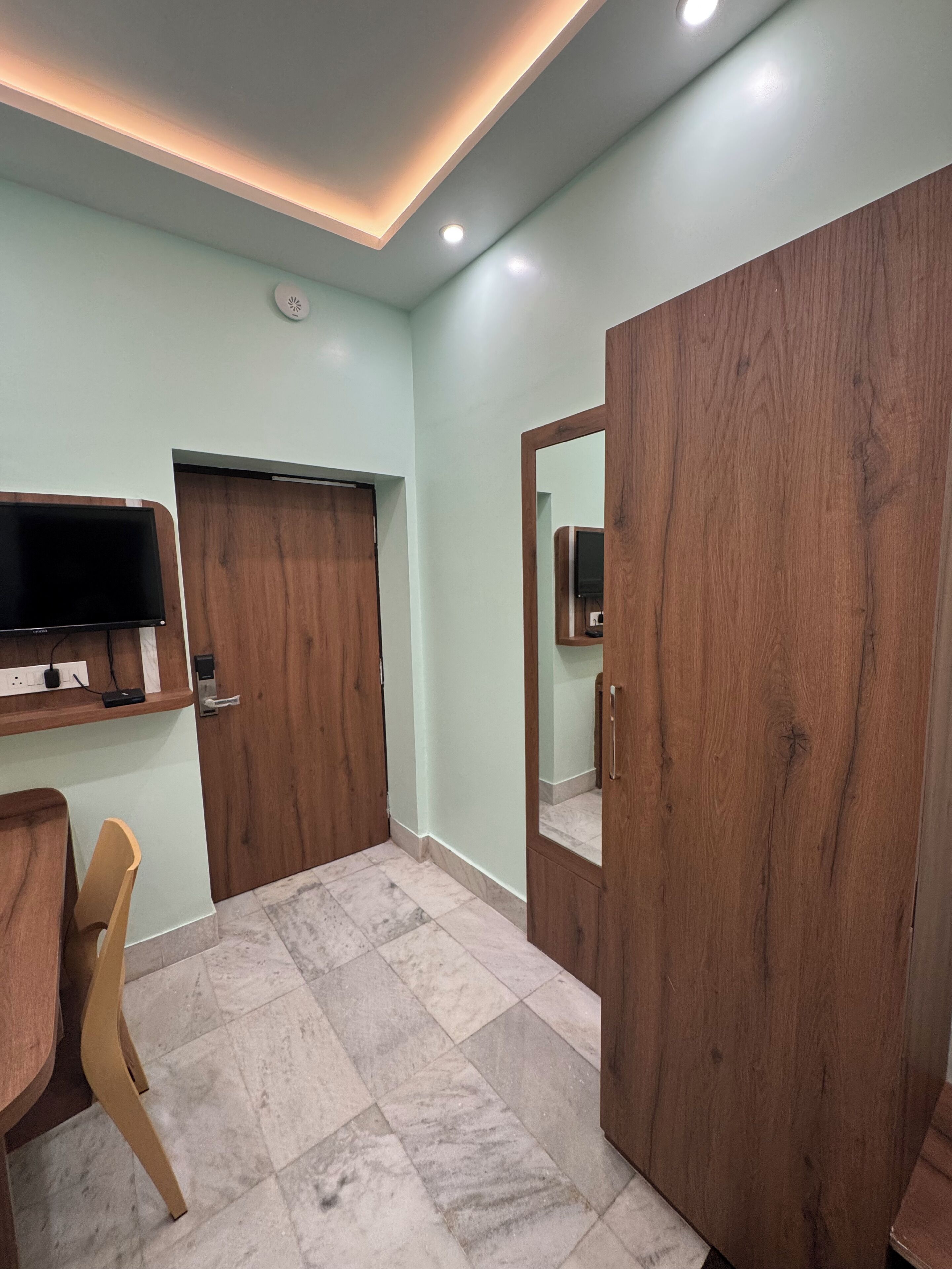 Una habitación con televisión montada en la pared, un armario de madera, una silla y piso de baldosas.